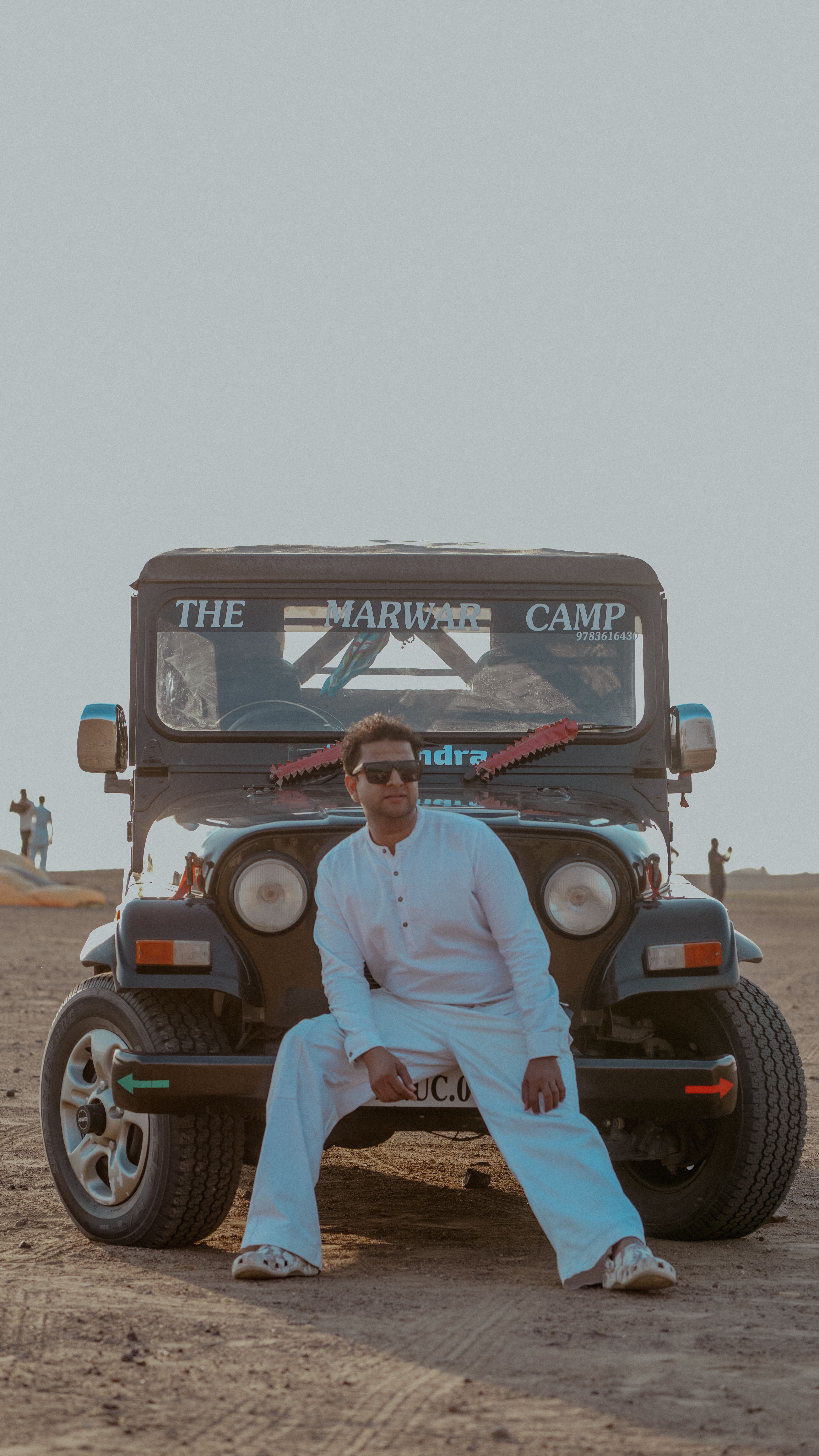 A man in white clothes sits on an SUV in the desert, exuding adventure and leisure.