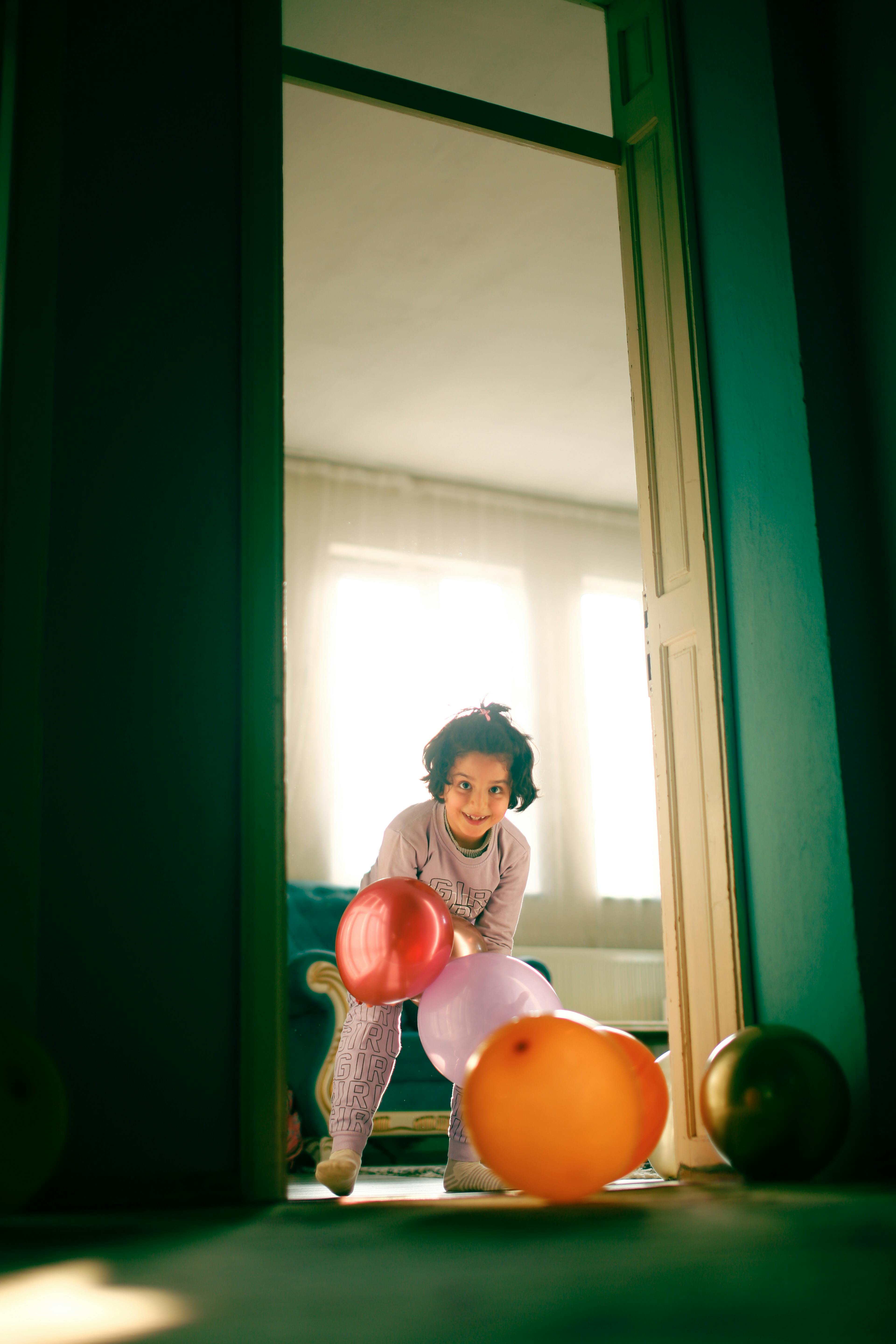 A cheerful girl stands in a doorway with colorful balloons, enjoying indoor playtime.