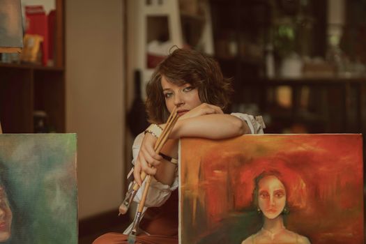 A woman artist in a studio with vibrant paintings, holding paintbrushes, showcasing creativity.