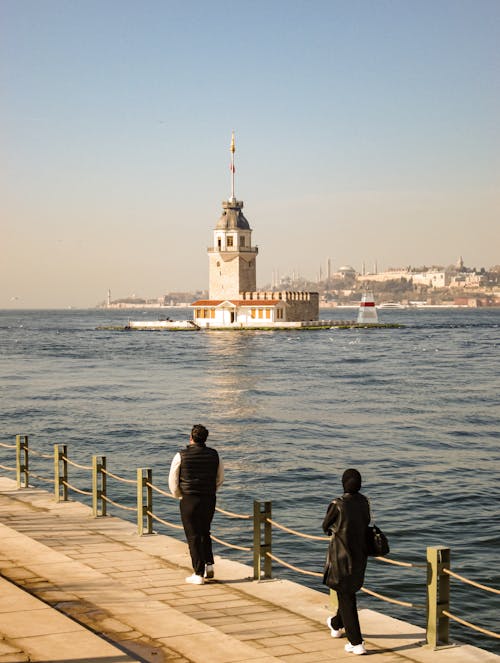 Two people walking along the water near a lighthouse