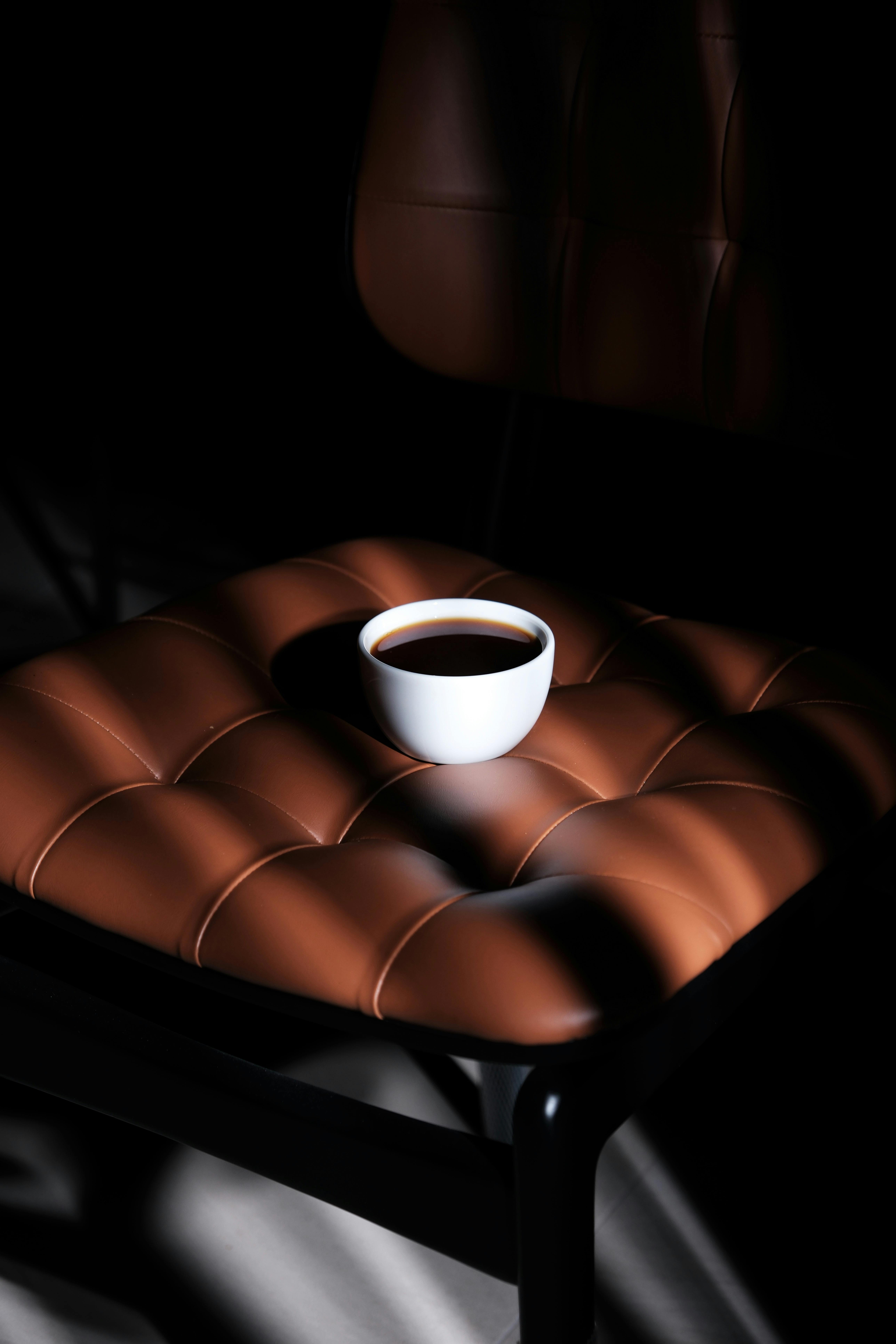 A white coffee cup sits on a luxurious leather chair, highlighted by dramatic lighting, creating an elegant scene.