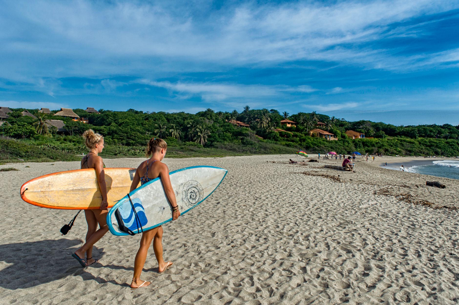 Two women carrying surfboards on a sandy tropical beach, with lush greenery in the background.