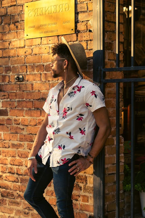 A man in a floral shirt and hat stands outside a brick building
