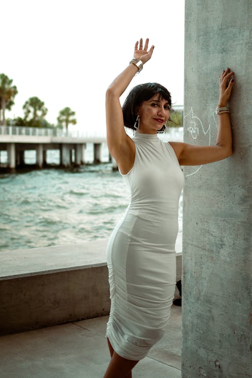 A woman in a white dress is posing near the water