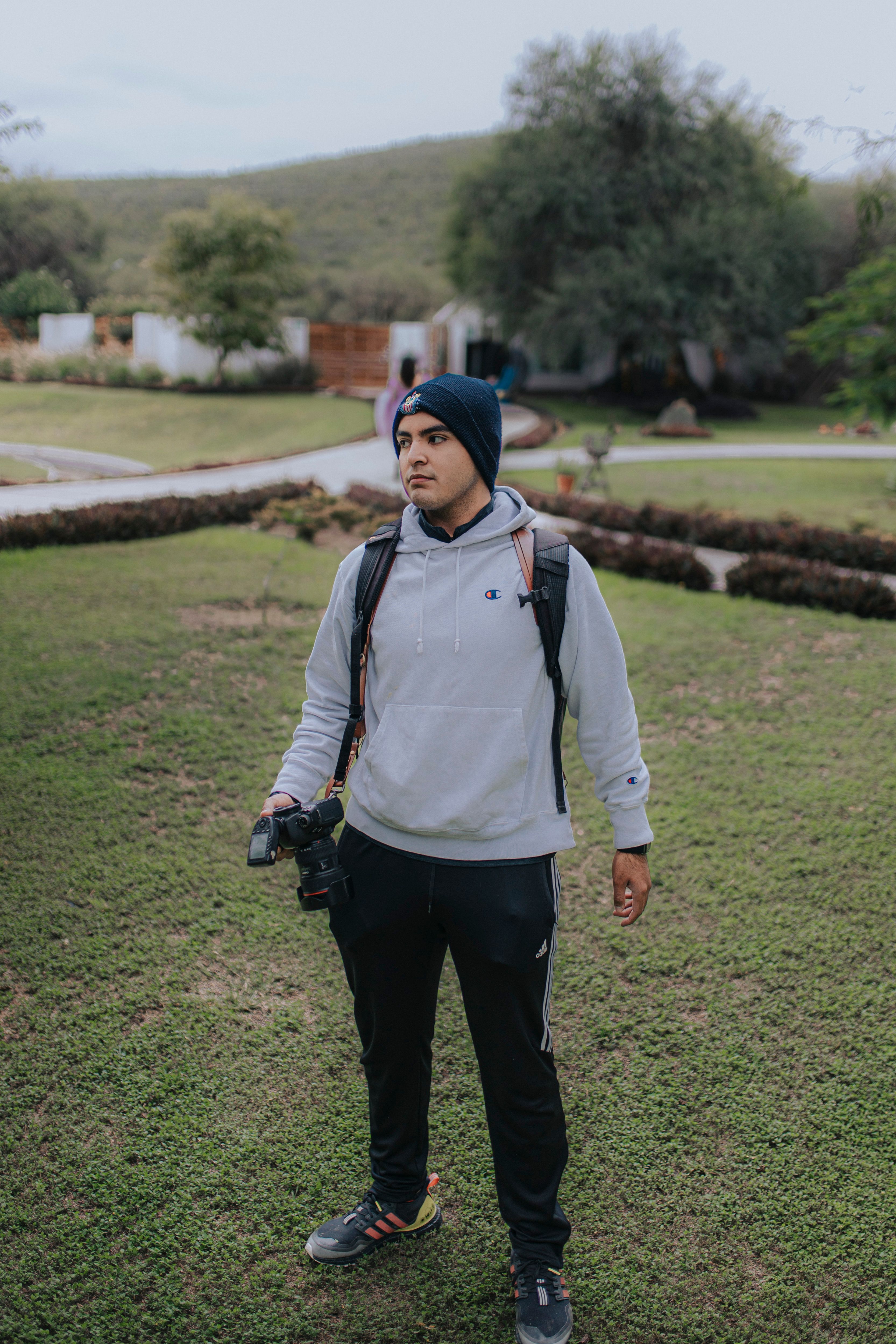 A young male photographer outdoors holding a digital camera, wearing a gray hoodie and knit hat.