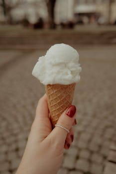 Close-up of a woman's hand with red nails holding ice cream cone on cobblestone street.