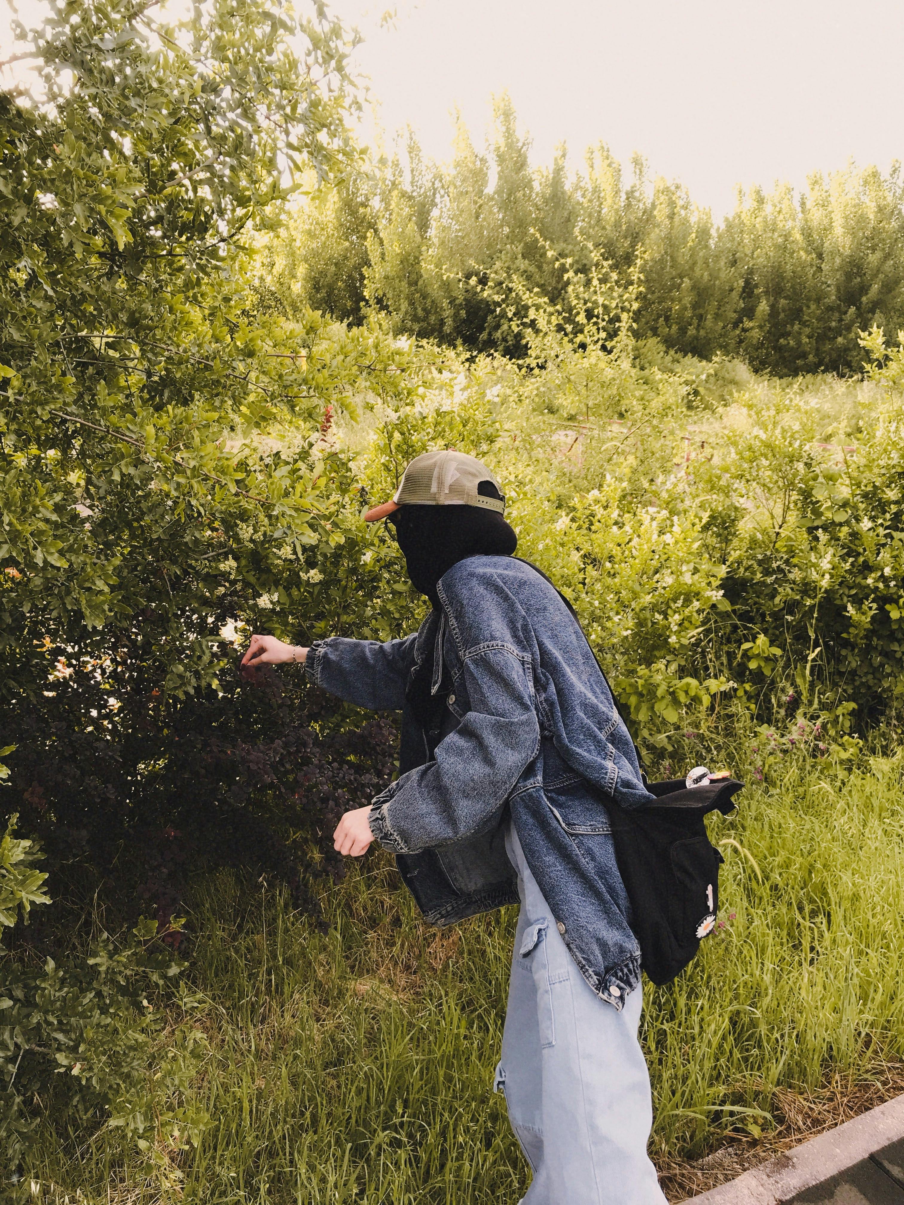 Woman in casual outfit picking leaves in a rural area, wearing a denim jacket and cap.