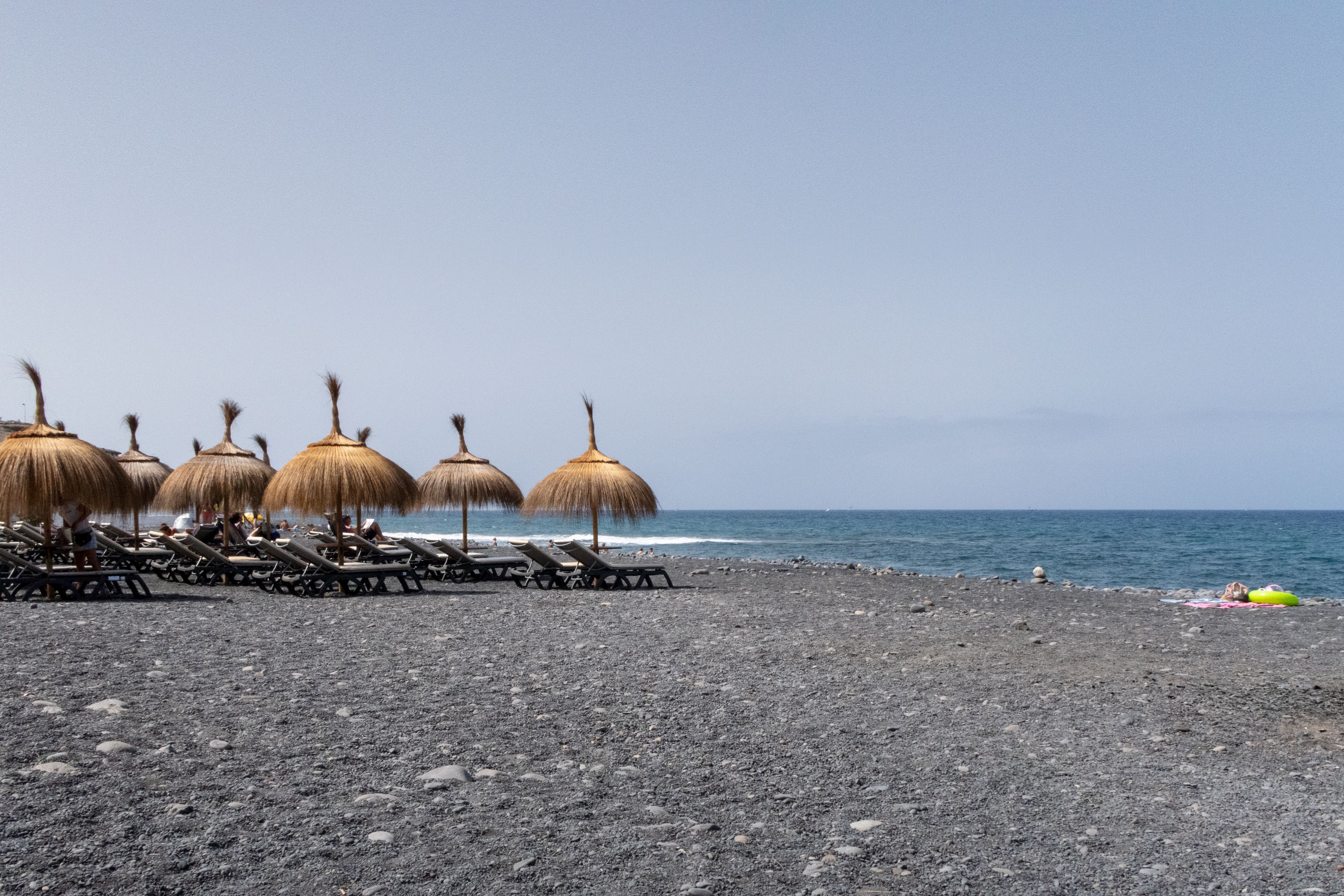 Peaceful beach setting in Lanzarote with straw umbrellas and deckchairs on a stone shore, under clear skies.
