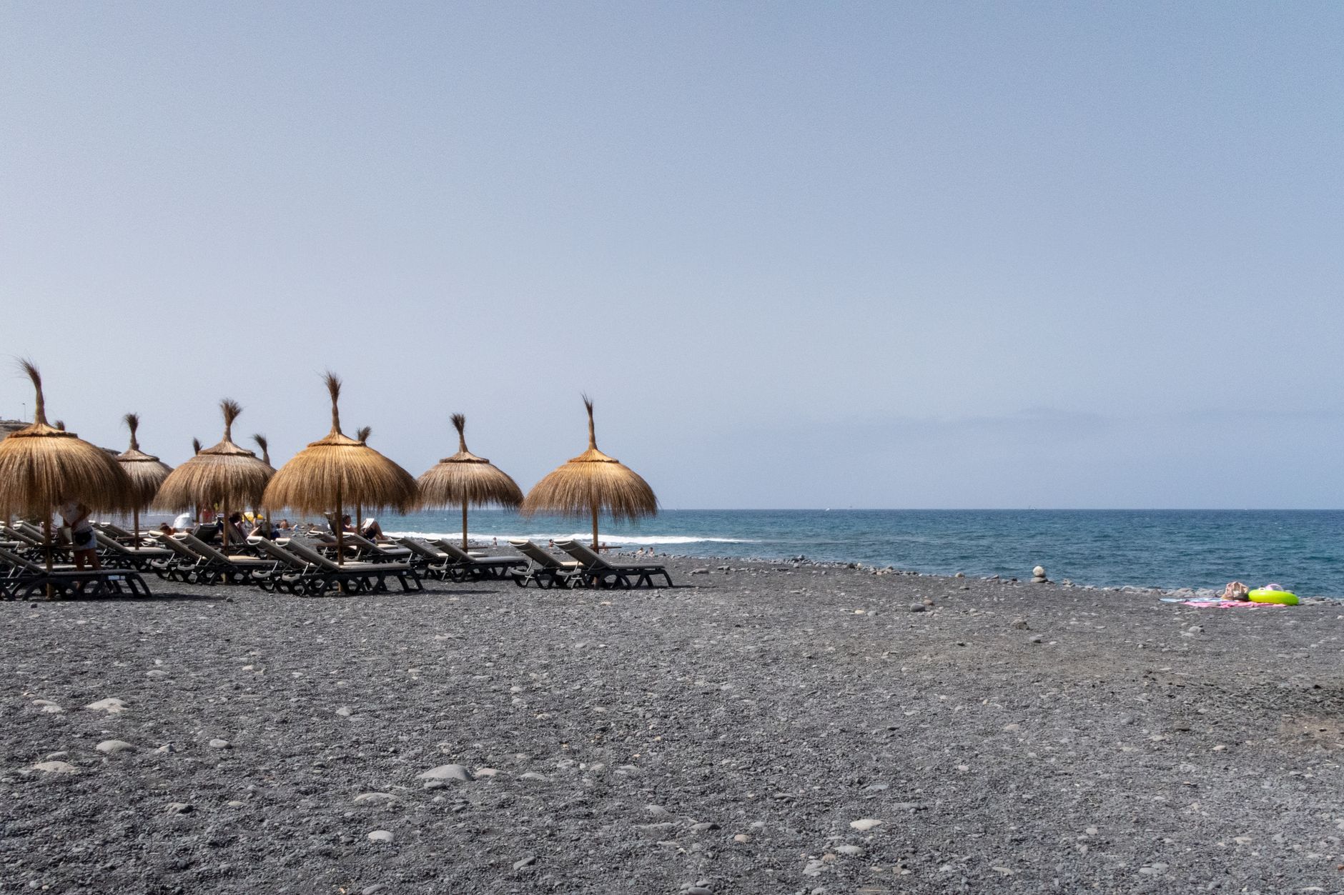 Peaceful beach setting in Lanzarote with straw umbrellas and deckchairs on a stone shore, under clear skies.