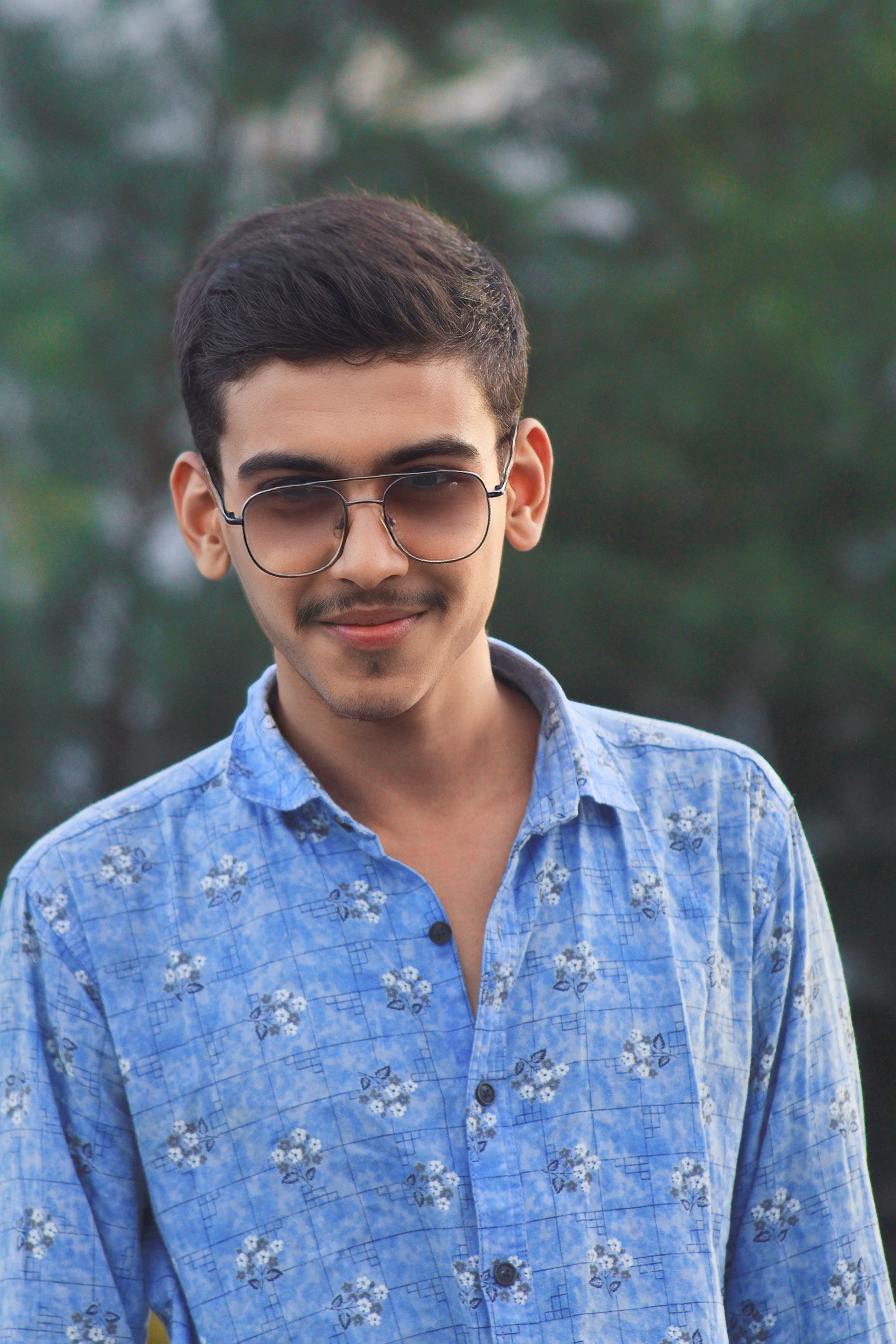 Smiling young man wearing sunglasses and a blue patterned shirt outdoors on a sunny day.