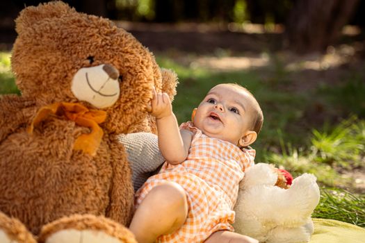 Cute baby in gingham romper enjoying time with teddy bears outdoors.