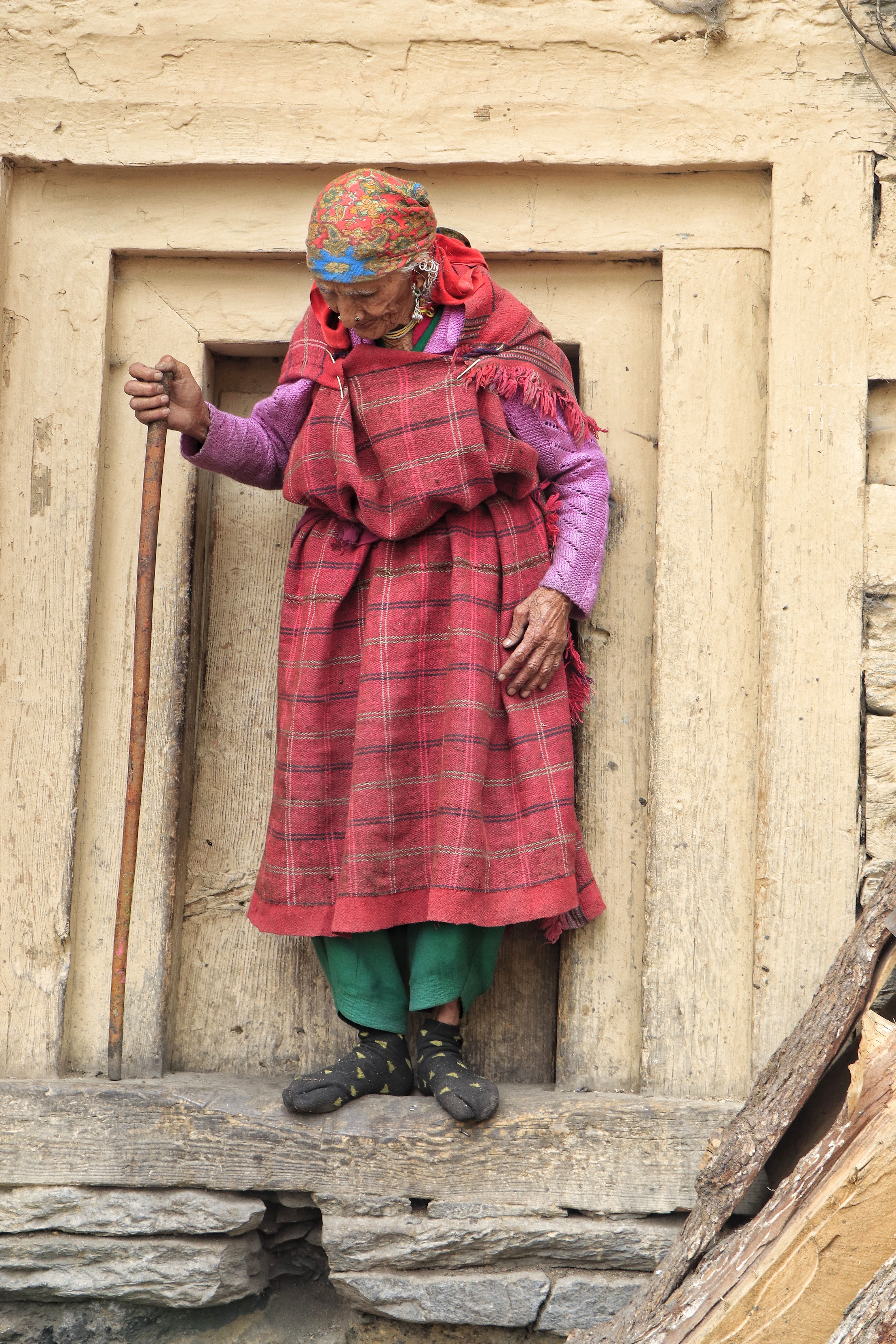 Elderly woman with headscarf leaning on a walking stick in rural setting.