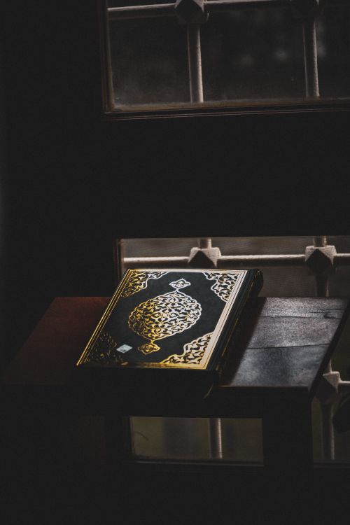 Free An ornate Quran resting on a wooden table near a window, capturing spiritual tranquility. Stock Photo