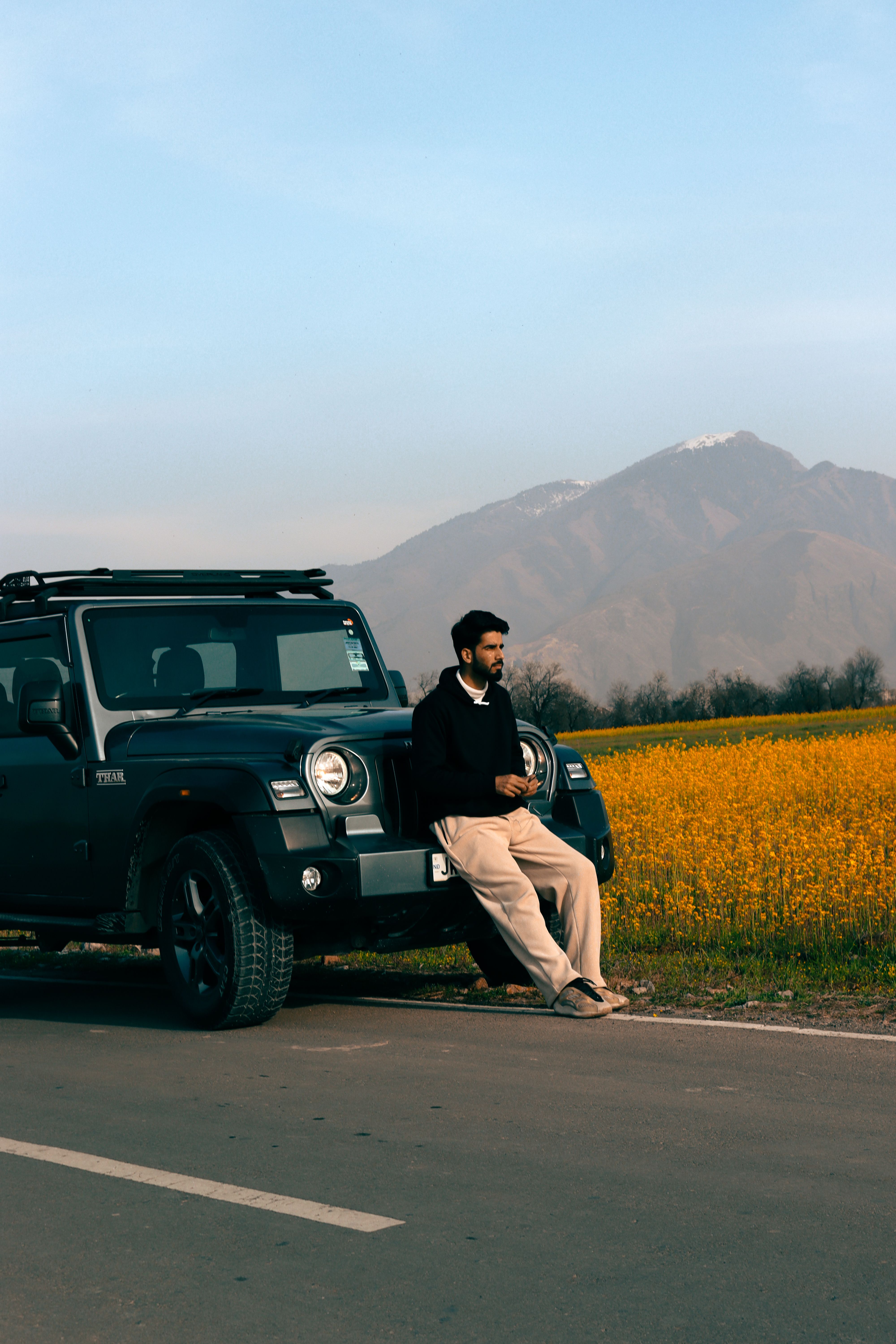 A man standing by a Mahindra Thar SUV with a mountain and yellow field backdrop, capturing adventure vibes.