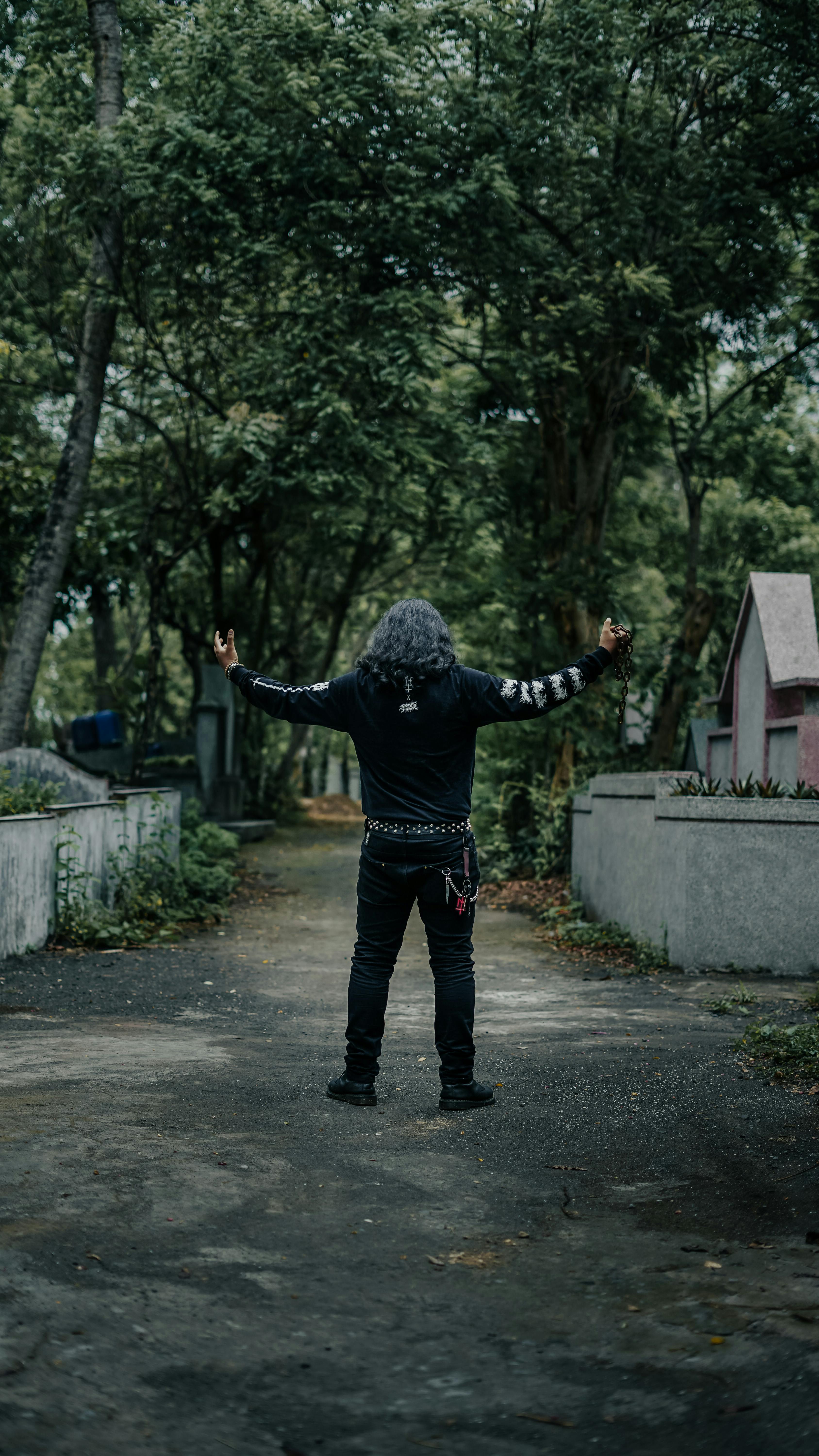 A man with long gray hair stands with arms raised in an outdoor forest path, surrounded by greenery.
