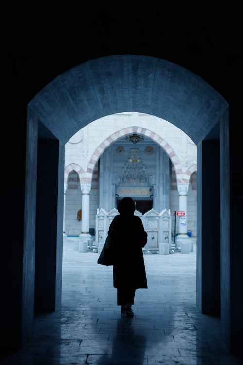 Free Silhouette of a woman standing in a mosque corridor, framed by an archway, highlighting Islamic architecture. Stock Photo