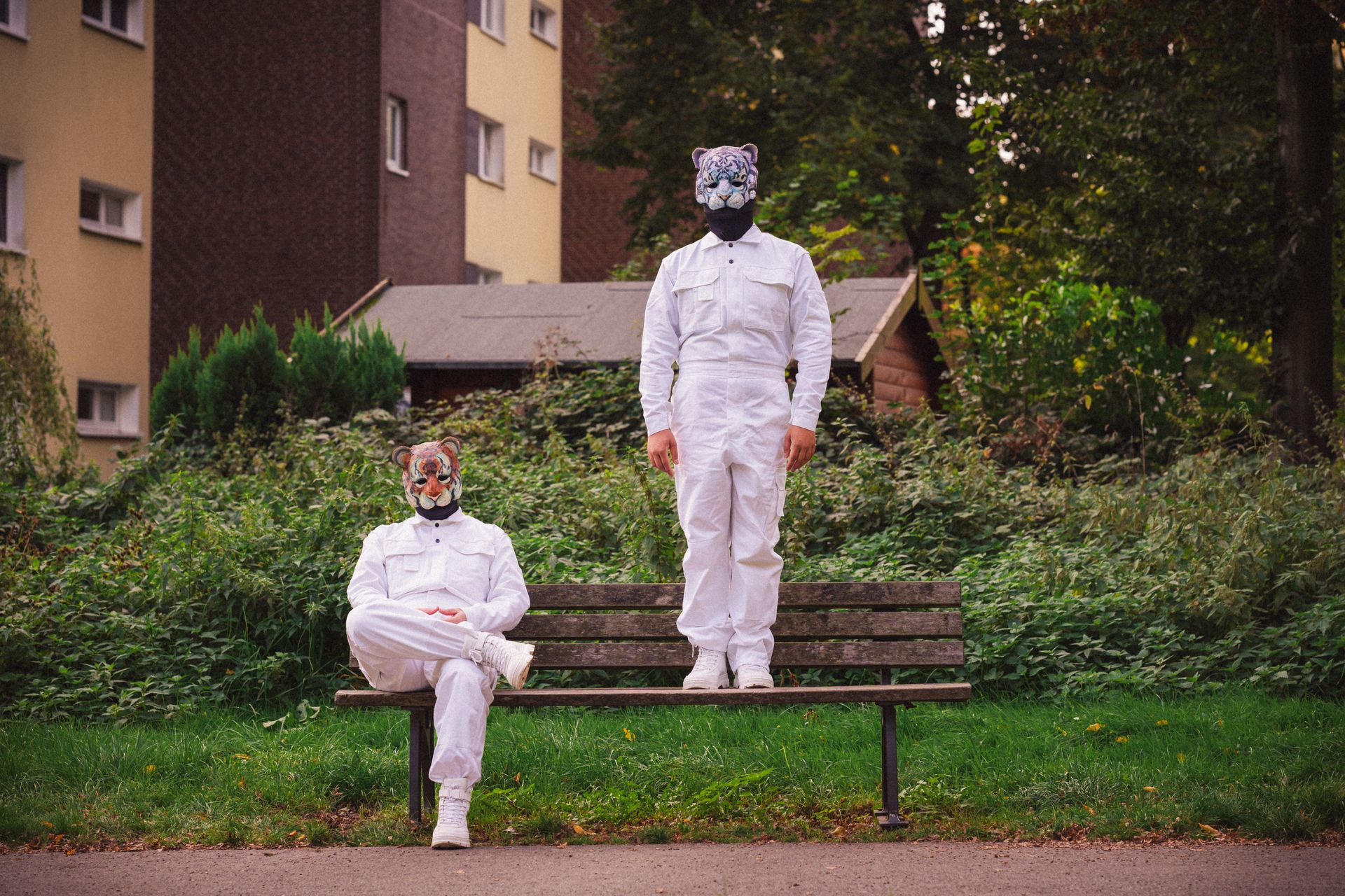 Photograph of two performers in animal masks and white outfits sitting and standing on a bench outdoors.
