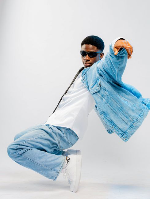 Fashionable black man in denim jacket and sunglasses striking a dynamic pose in a studio setting.
