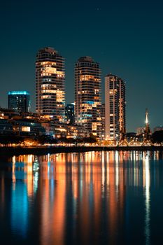 Vibrant night cityscape with illuminated skyscrapers reflecting on water, creating a serene urban view.