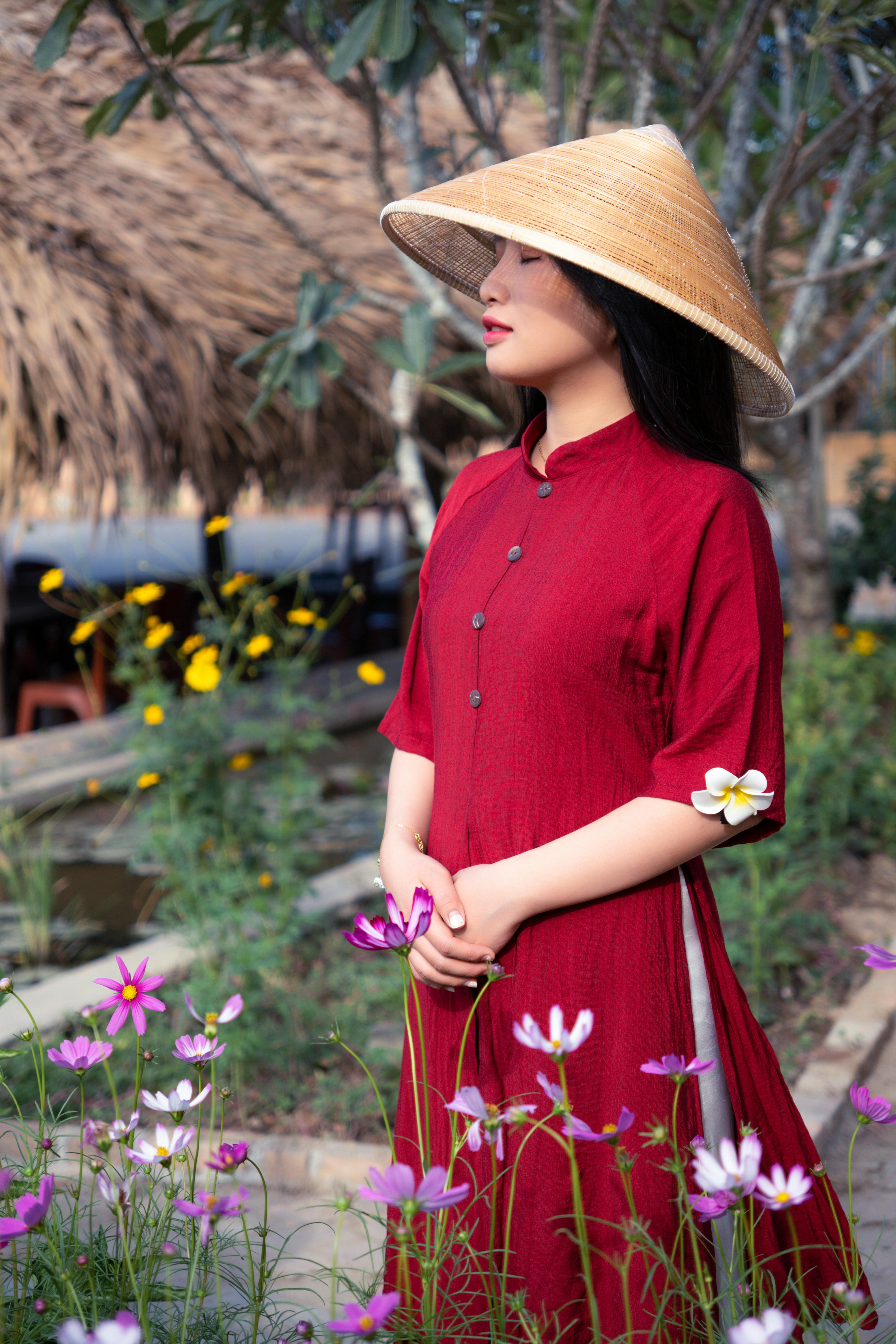 Asian woman in traditional red dress and conical hat standing amidst vibrant flowers.
