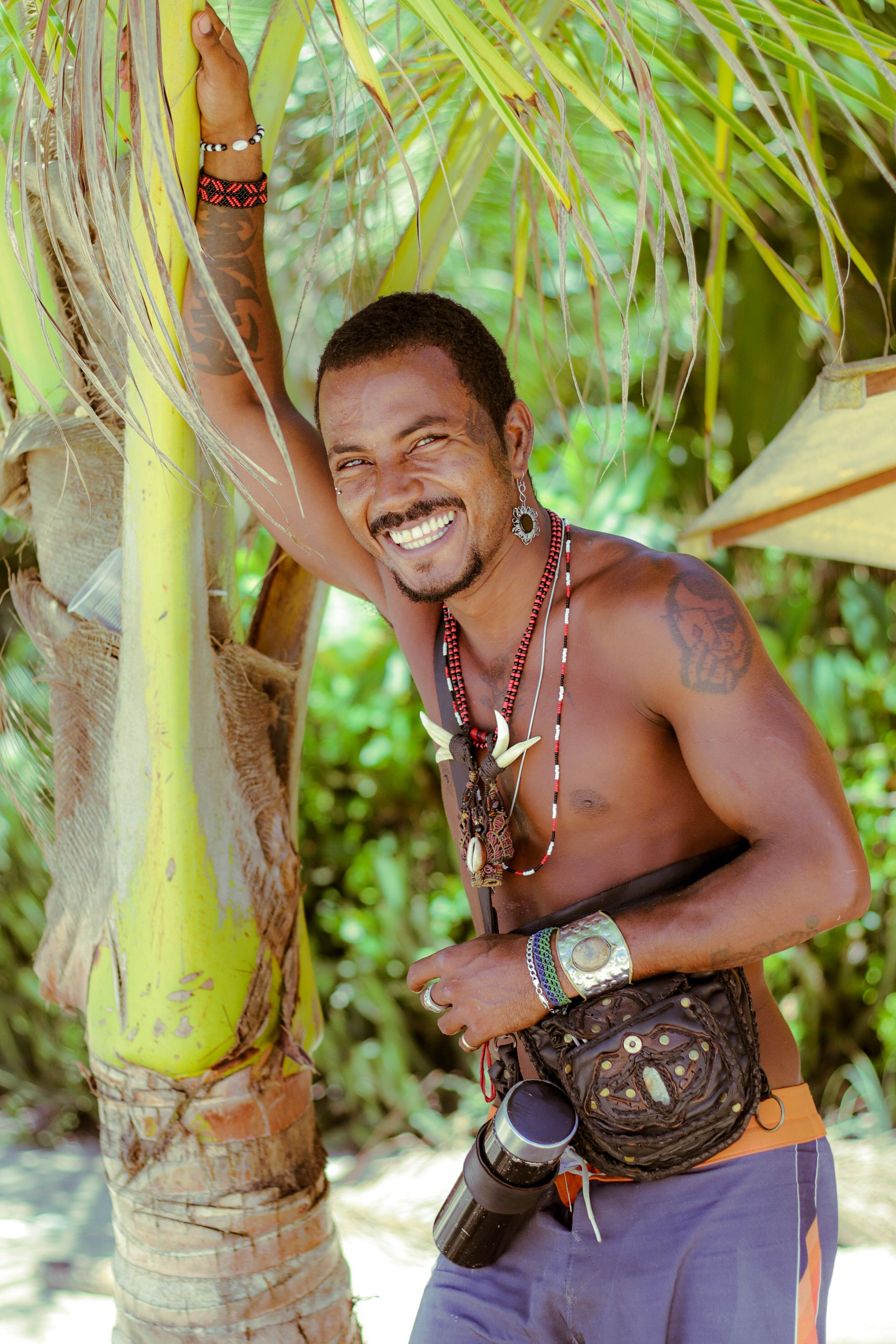 Topless man with tattoos and accessories smiling by a palm tree in tropical setting.