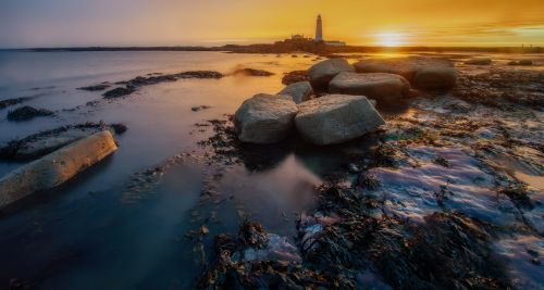 Free Serene view of lighthouse at sunset with rocks and calm sea reflecting warm hues. Stock Photo