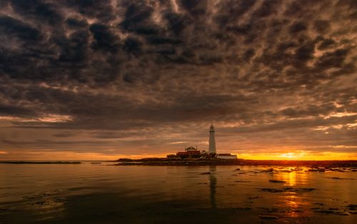 Free A lighthouse by the sea at sunset, surrounded by dramatic clouds and rocky shores. Stock Photo