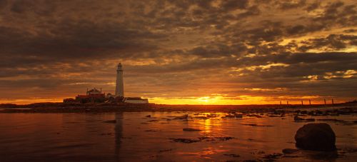 Free A dramatic sunset over the coast featuring a lighthouse, reflecting vibrant colors in the sea. Stock Photo
