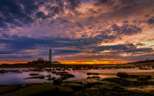 Free Stunning sunset sky over a lighthouse by the rocky coast, reflecting vibrant colors on sea and rocks. Stock Photo