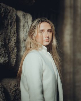A blonde woman poses stylishly by an ancient stone wall, exuding elegance and grace.