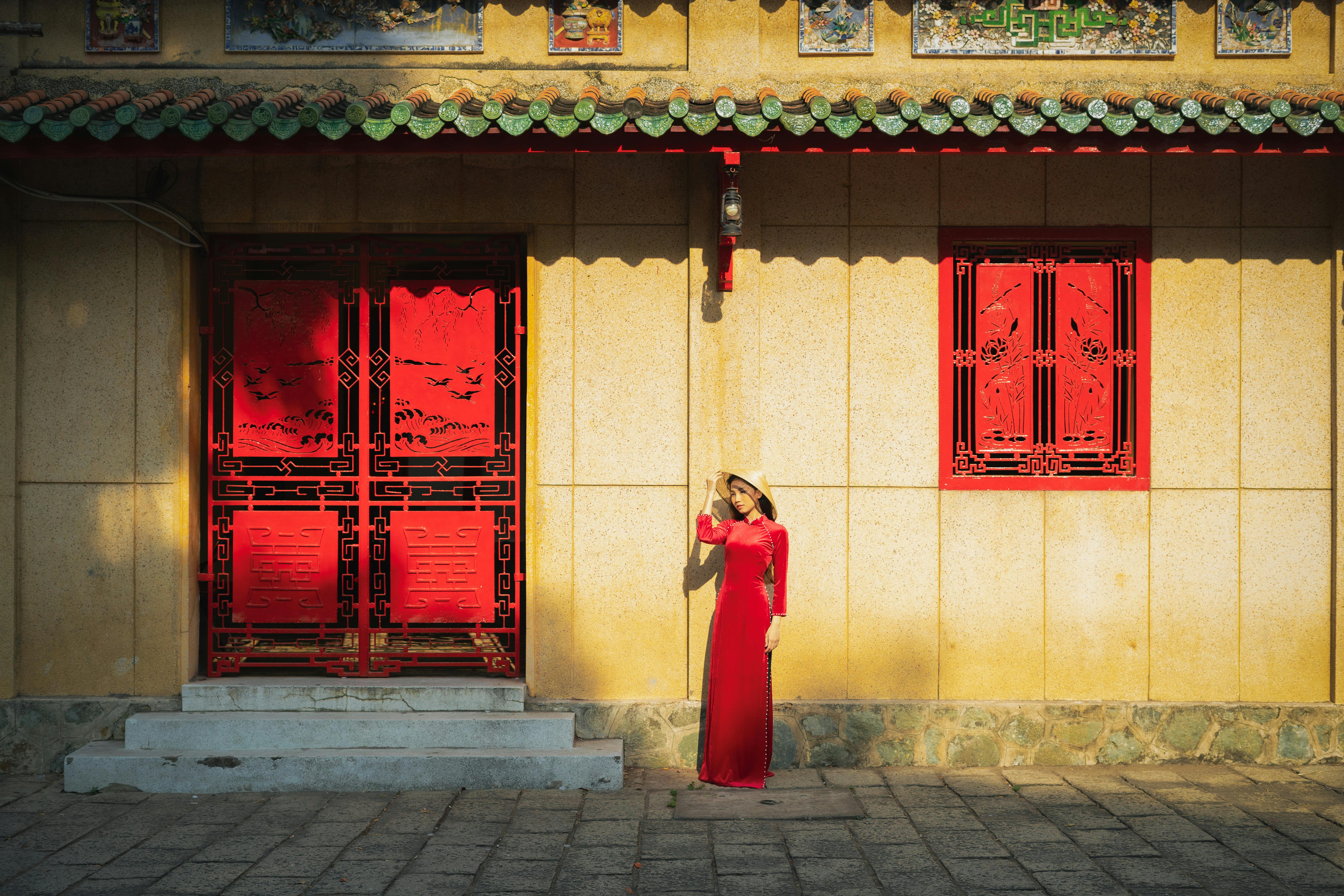 Asian woman in traditional red dress standing by ornate architecture in city.