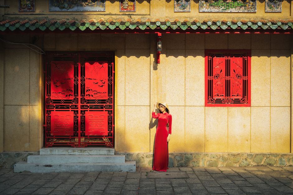 Asian woman in traditional red dress standing by ornate architecture in city.