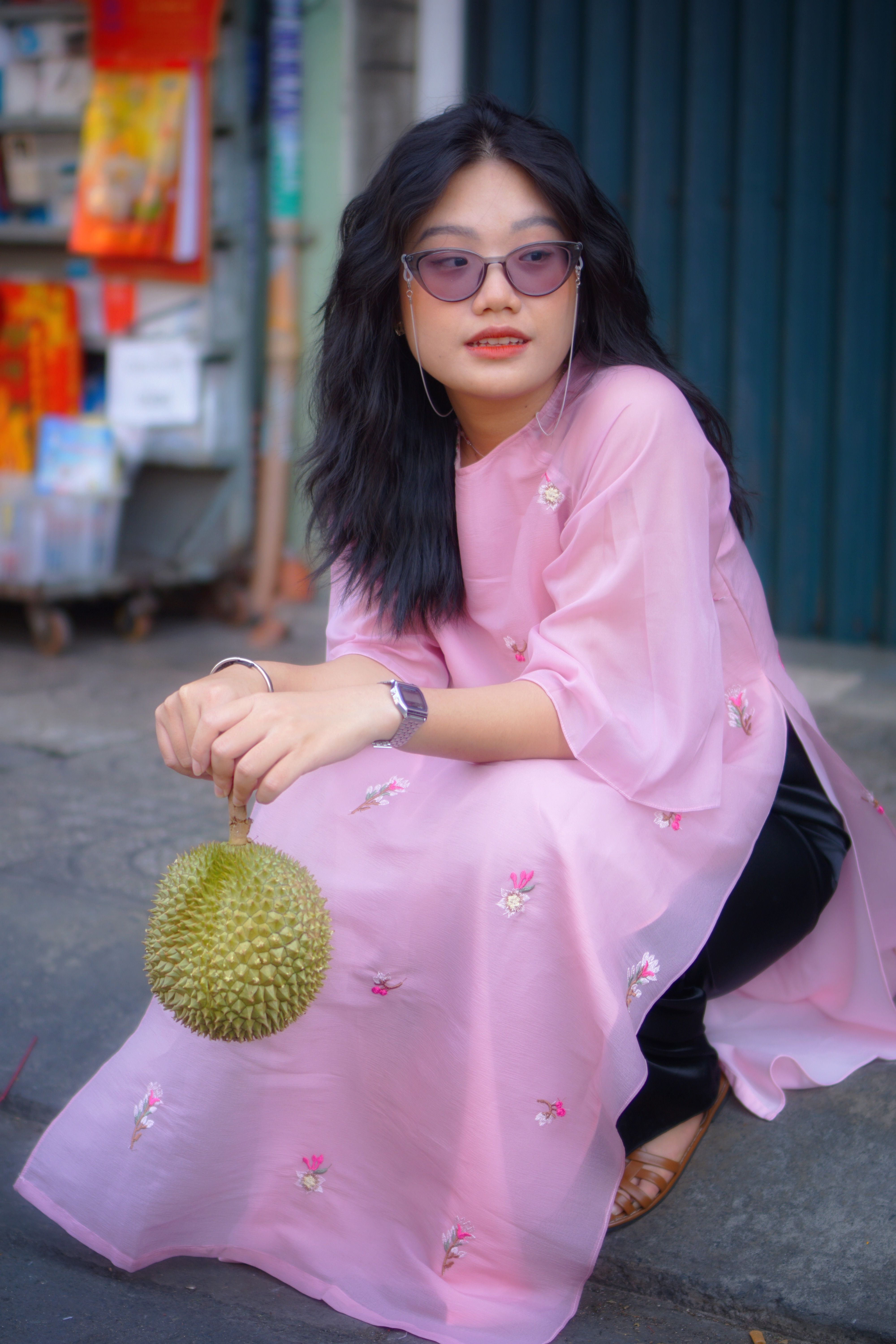 Stylish woman in a pink dress with sunglasses holding a durian in an urban setting.