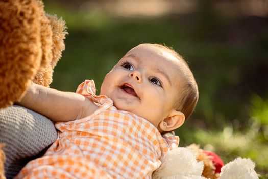 Charming baby in checkered dress outdoors, enjoying playtime with her teddy bear.