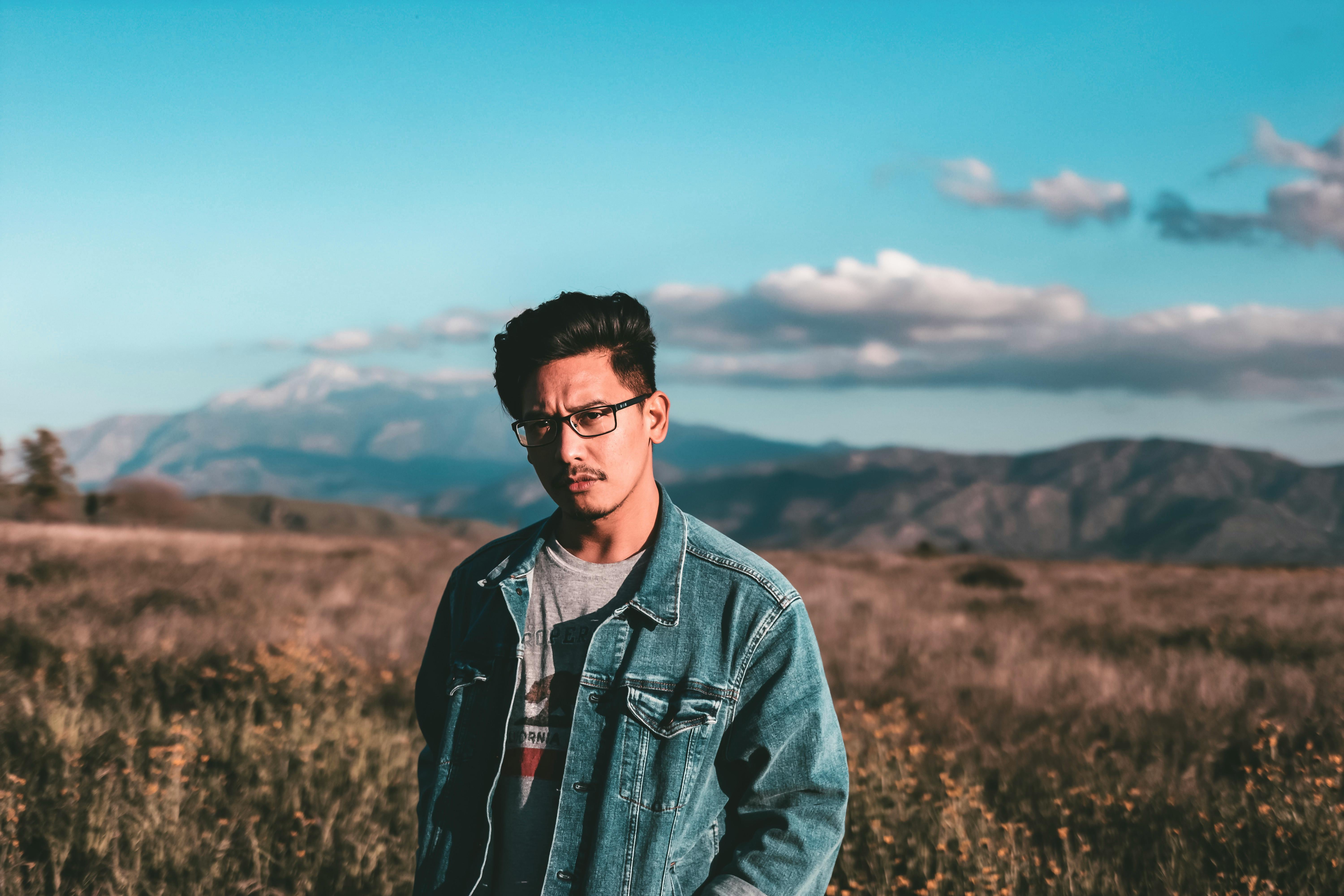 一名身穿牛仔夹克的亚洲男子，站在风景优美的山地景观中，背后是蓝天