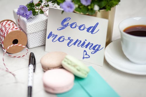 Free Filled Teacup With Saucer Beside Good Morning Card and Pen and Macaroons Stock Photo