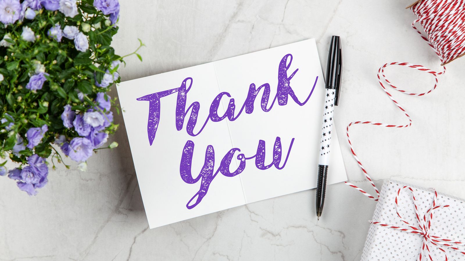 A thank-you card, purple flowers, a pen and wrapped gift on a light surface