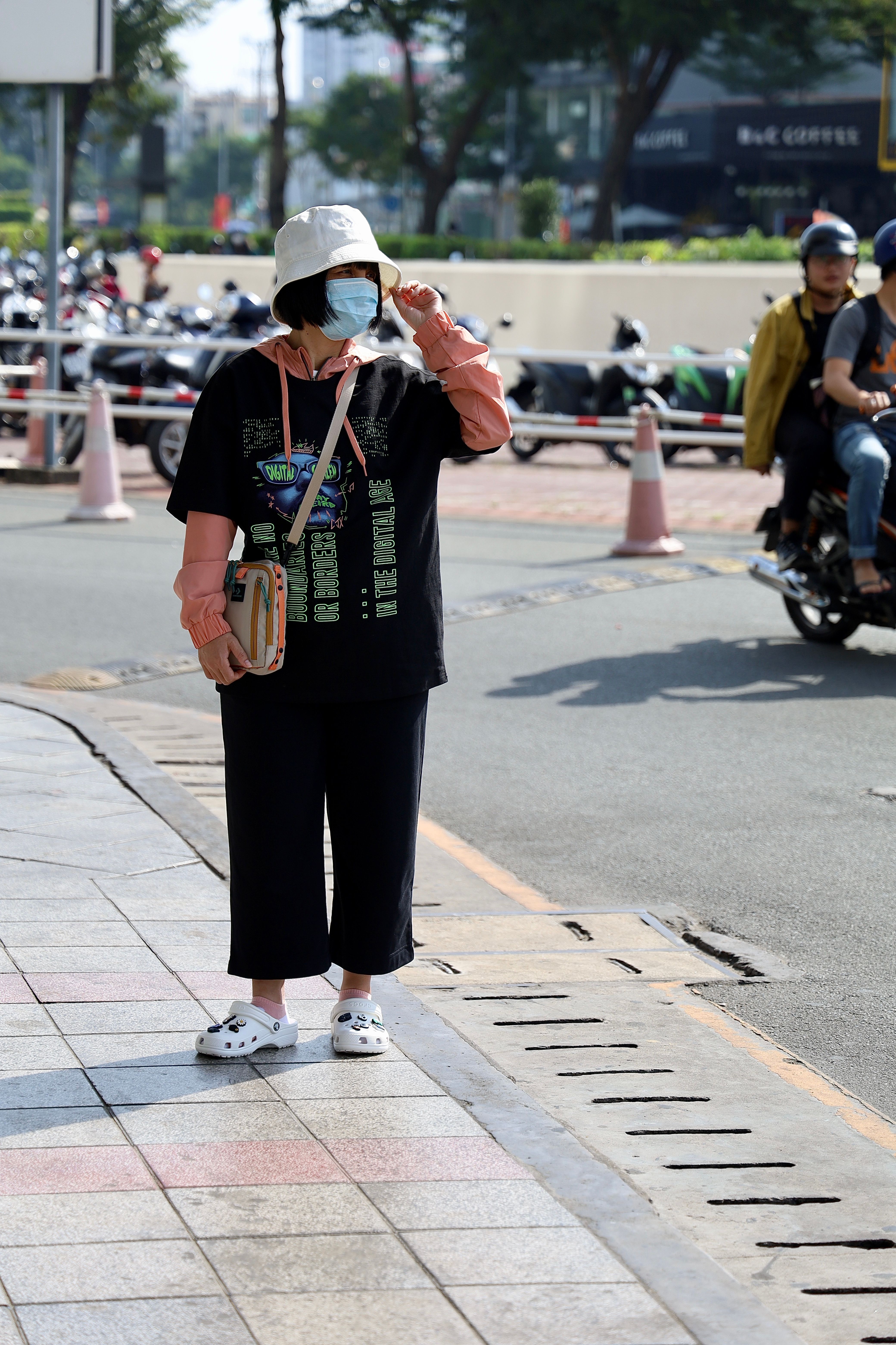 Woman with hat and mask standing on a city street sidewalk in Ho Chi Minh City, Vietnam.