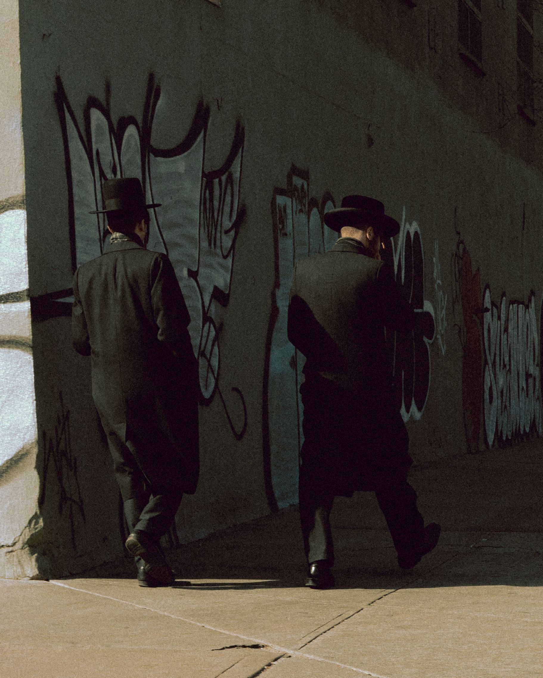 Two men in black coats walk past graffiti-covered walls on an urban sidewalk.