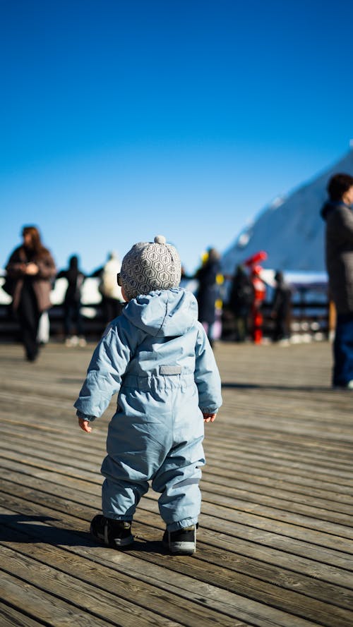 A baby in a blue jacket walking on a wooden pier