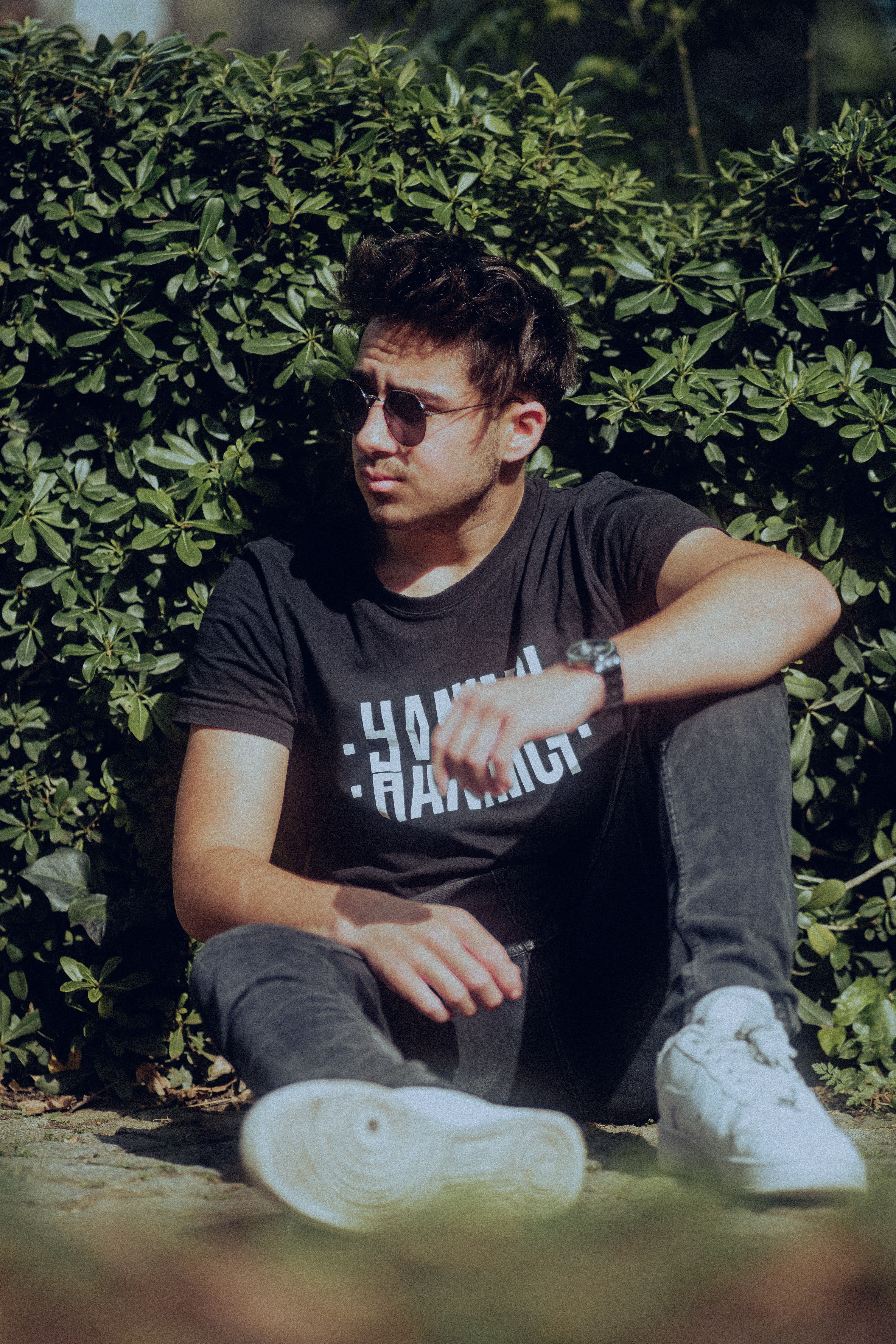 Young man wearing sunglasses and casual outfit, sitting against a hedge in an urban setting.