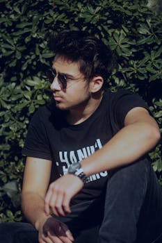 Stylish young man in sunglasses sitting against leafy background on a sunny day.