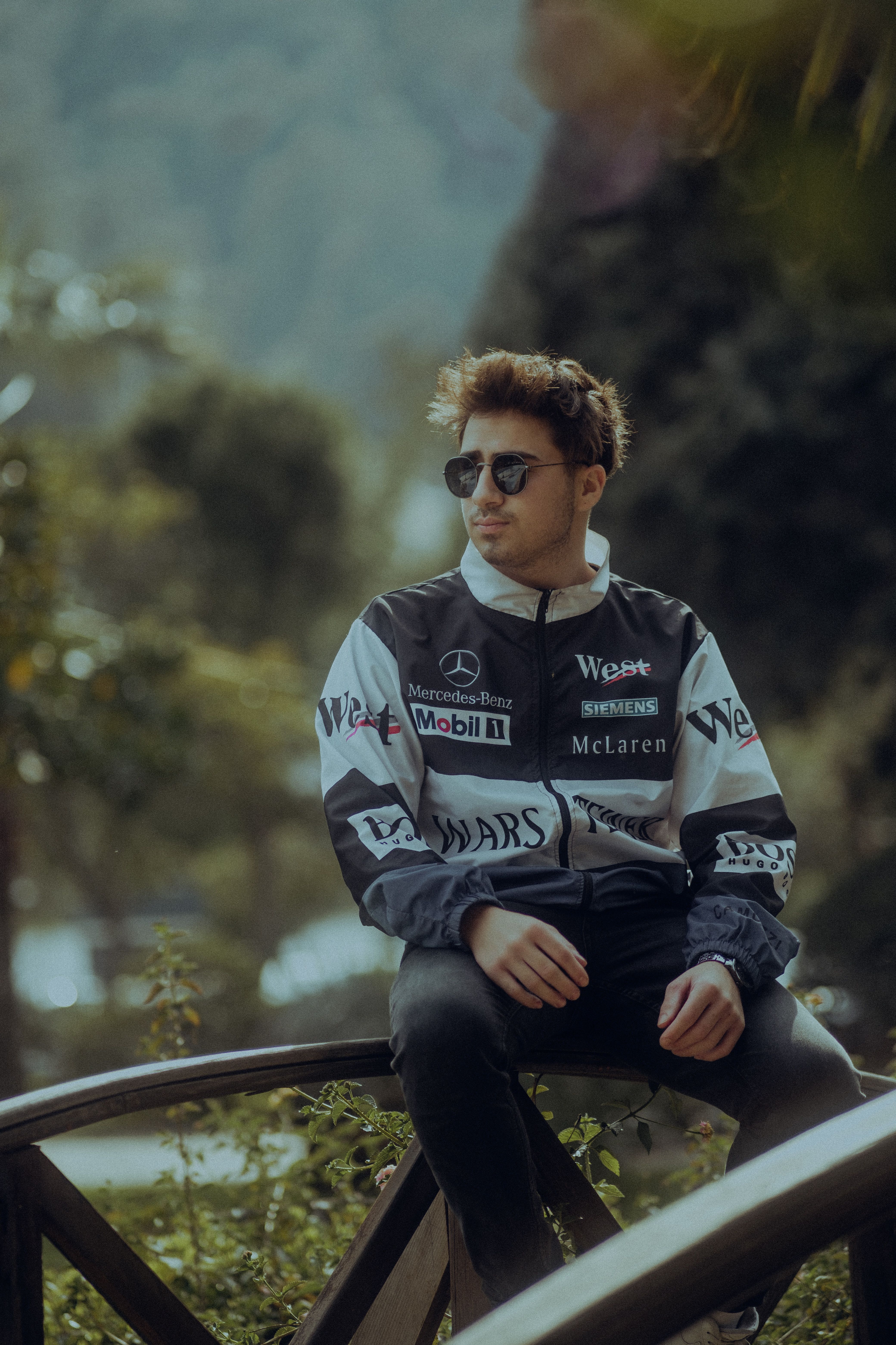 Stylish man wearing a racing jacket and sunglasses outdoors, exuding a confident and relaxed vibe.