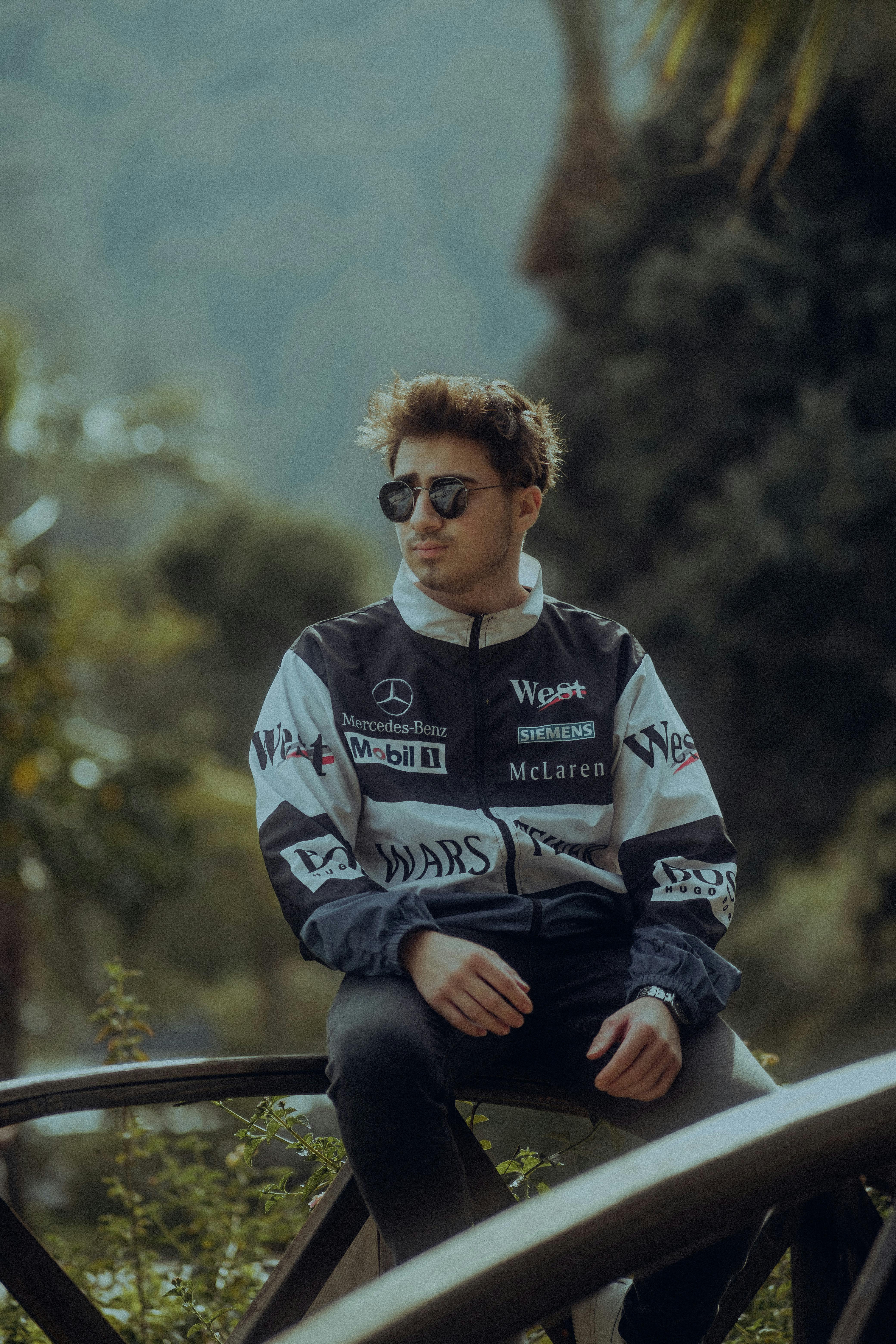 A young man in a printed jacket and sunglasses sitting casually on a footbridge outdoors.