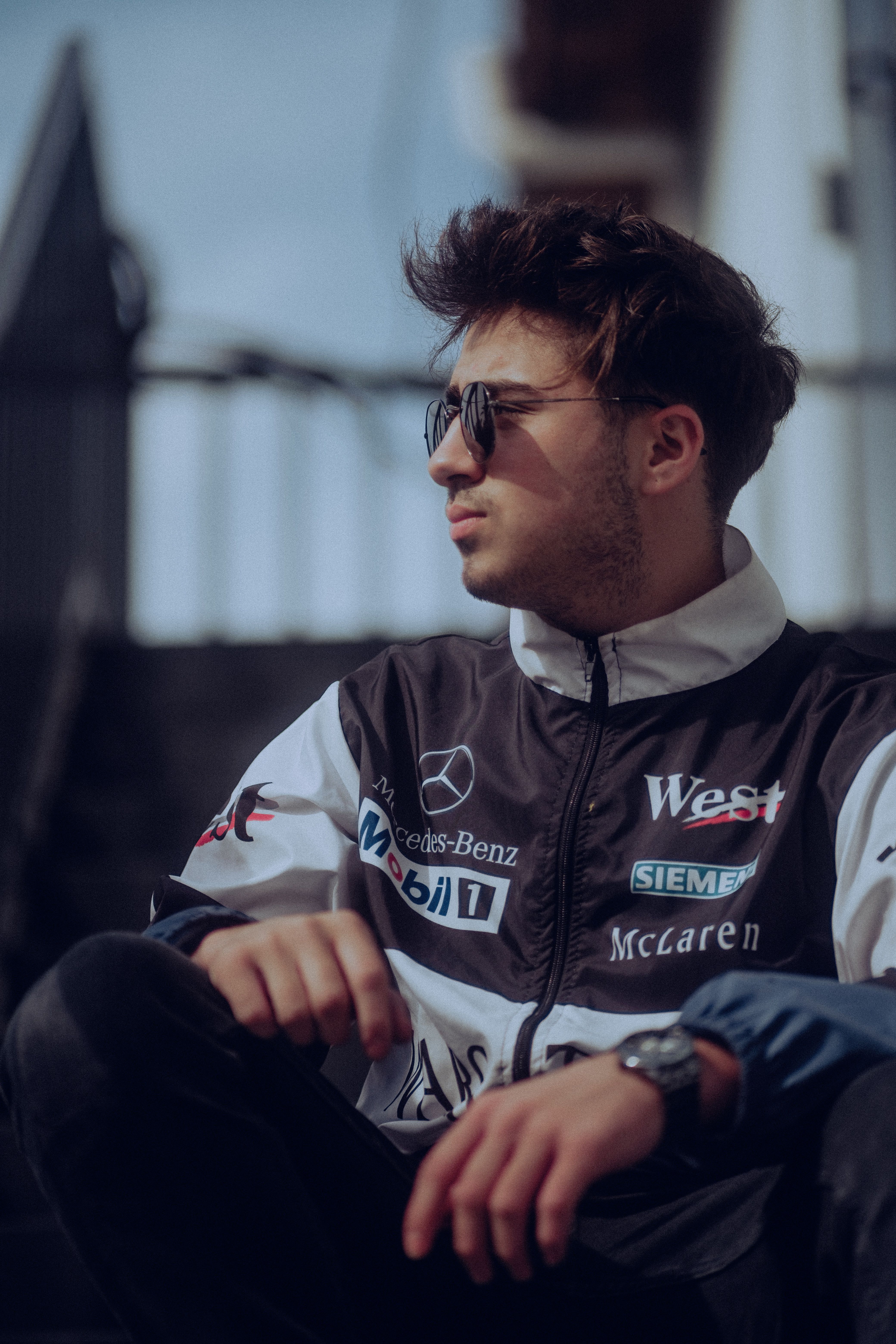 Fashionable man in a racing jacket sits outdoors wearing sunglasses, exuding urban style.