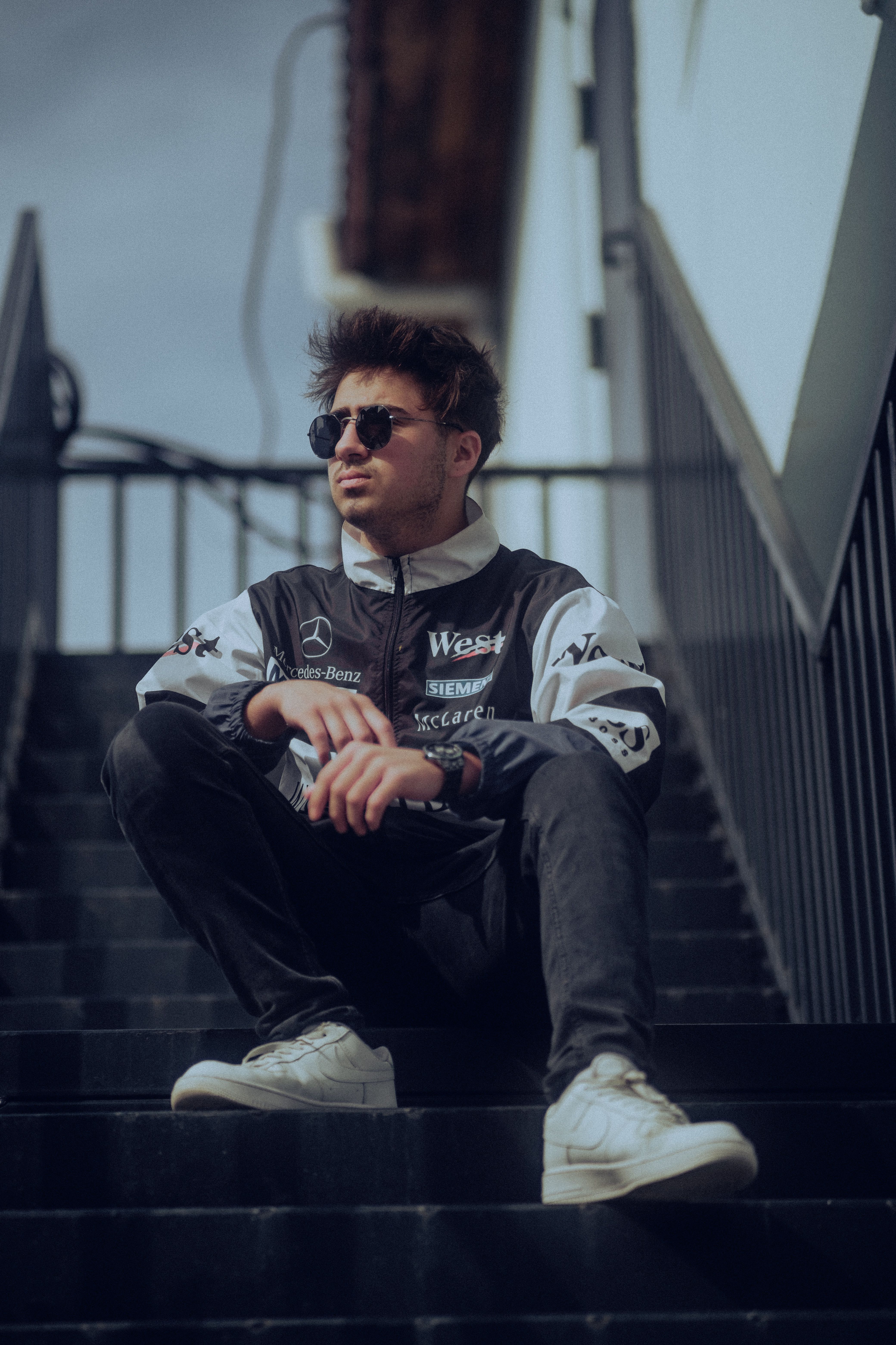 Stylish man in sunglasses and jacket sitting on stairs outdoors.