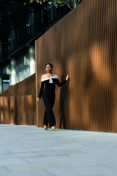A woman in a black jumpsuit is standing by a wall