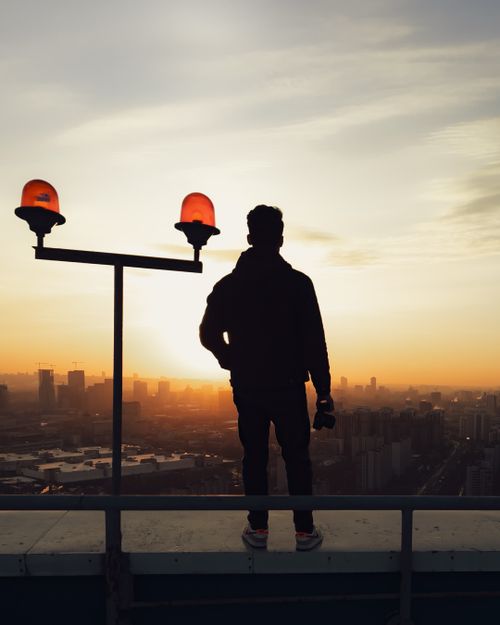 Free A man stands on a rooftop overlooking a cityscape, silhouetted against a dramatic sunset. Stock Photo
