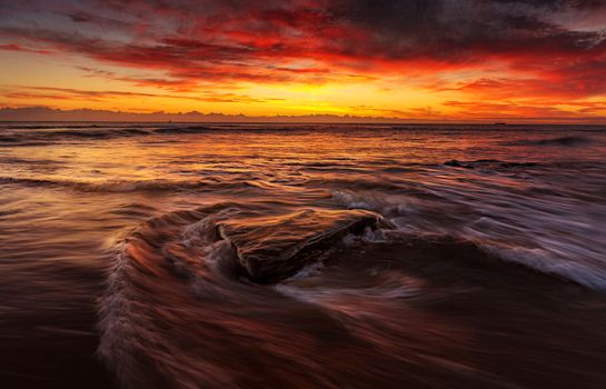A dramatic sunset with red skies and ocean waves crashing over rocks.