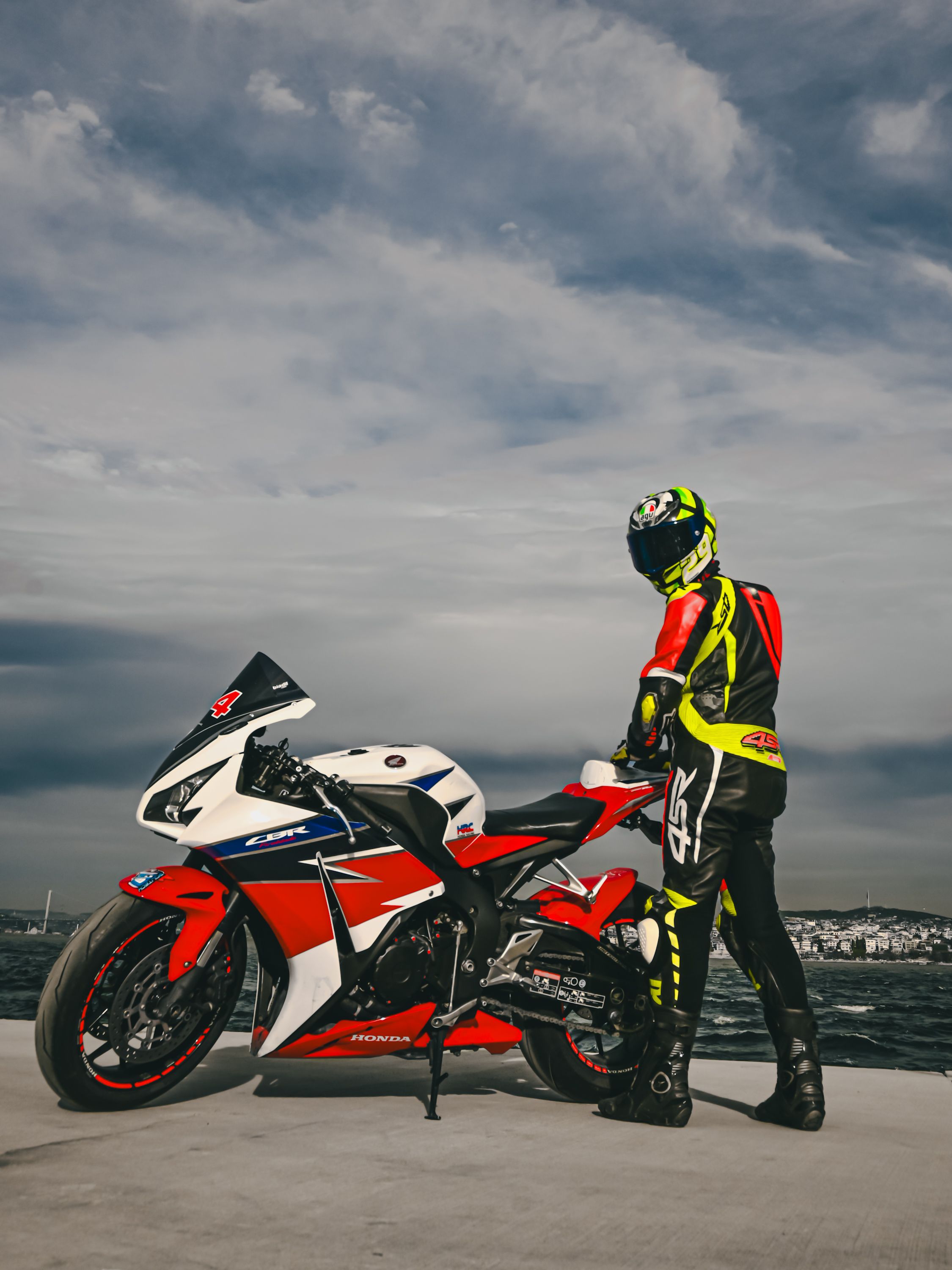 A motorcyclist in a leather suit stands by a racing motorcycle on a promenade near the sea.
